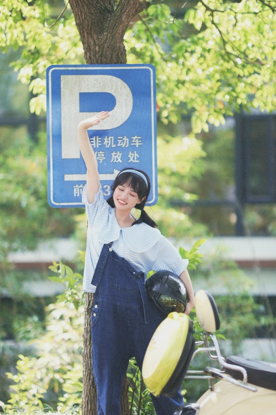 一位年轻女子手持蓝色标志站在一棵树旁并停着一辆摩托车。