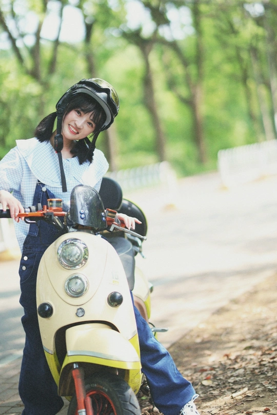 一个戴头盔的年轻女子站在一个小女孩旁边 小女孩坐在一辆小摩托车上。