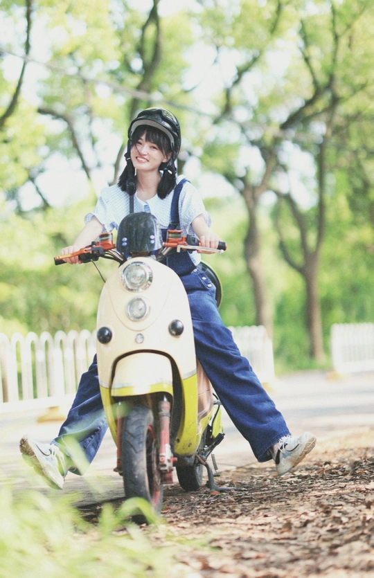 一个戴头盔的年轻女孩和男孩骑着小滑板车沿着树木成荫的道路向下行驶。