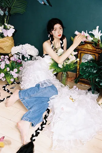 一个穿着白色连衣裙的年轻女子坐在地板上 周围环绕着植物和鲜花。