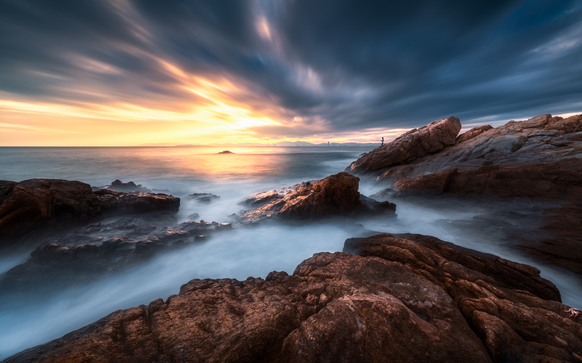 夕阳或日出时 岩石海滩的长时间曝光照片 太阳位于天空中的云层上方 周围有水域。