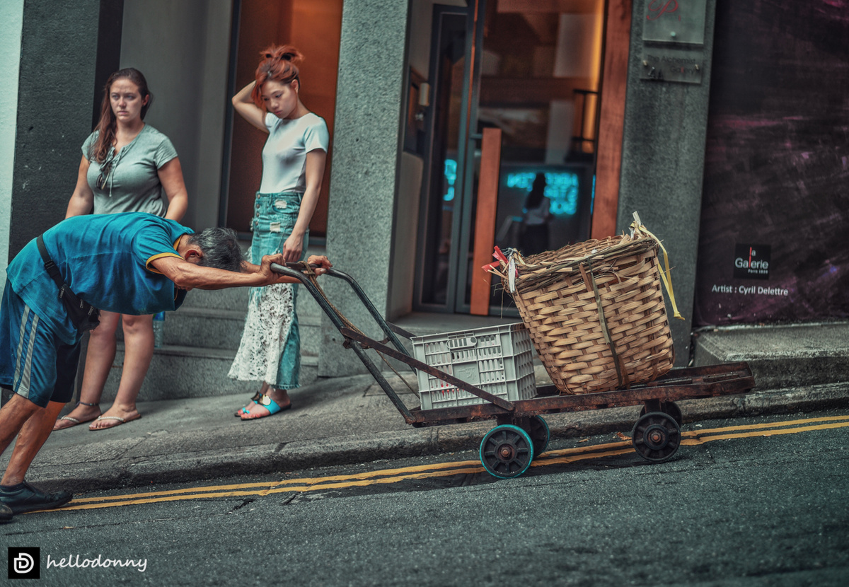 一个男子推着一辆小车沿街而下 与此同时 一个女子推着一篮子东西沿人行道而行。