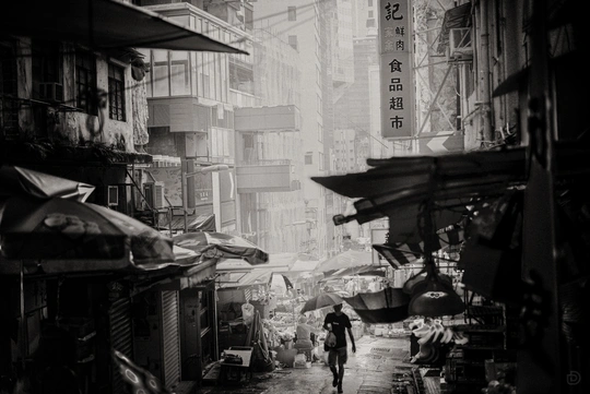 一张黑白照片 展示了一个亚洲城市的巷子 人们手拿雨伞走在街道上。