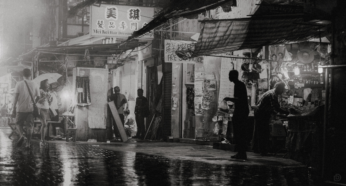 一张黑白照片 描绘了人们在雨夜沿着湿漉漉的巷子行走。
