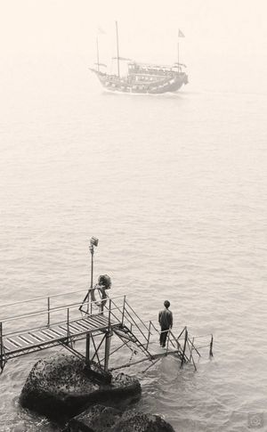 一张黑白照片 一个男子和一个男孩站在码头上 眺望着海洋 远处有一艘船 还有一个人正在桥上走。