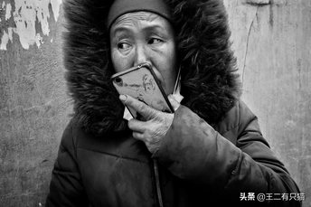 一张黑白照片 一位老人戴着帽子 披着斗篷 拿着手机对着嘴