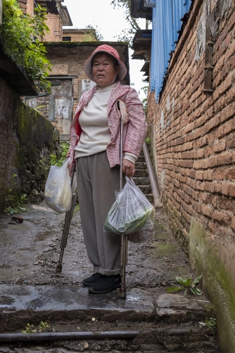 一位老太太沿着一条狭窄的巷子走下来 手里拿着一根木棍和一只装满杂货的塑料袋 站在楼梯上。