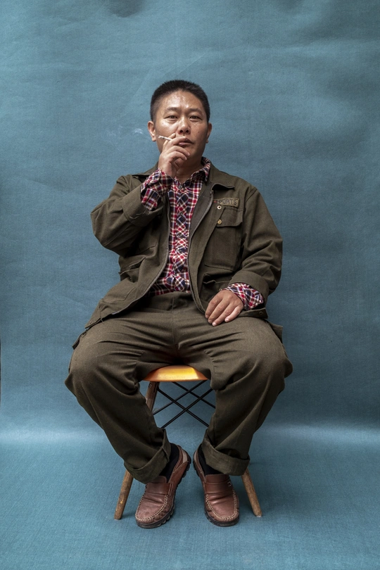 一个穿着棕色西装的男子坐在蓝色的椅子上 双手放在木质的矮凳上。