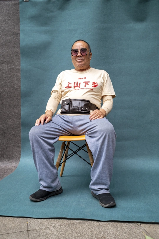 一个戴太阳镜和一件带有摄像头的衬衫的男人坐在蓝色的背景板前一把椅子上