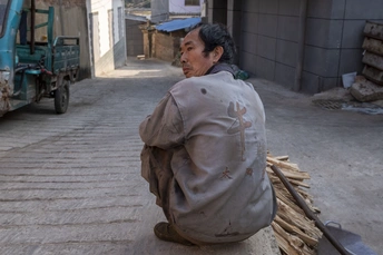 一个老人坐在街边 旁边有一堆木头和一辆卡车。