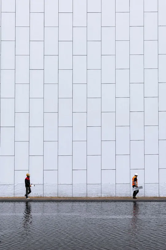 一个人打着伞走在一条街道上 前面有一座大型白色建筑 建筑前面有水。