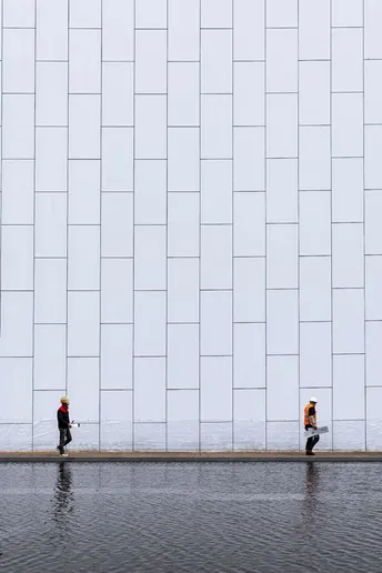 一个人打着伞走在一条街道上 前面有一座大型白色建筑 建筑前面有水。