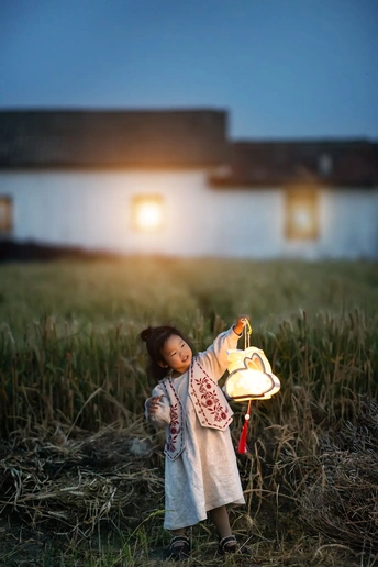 一个小女孩拿着一个小的灯笼 里面点着蜡烛 站在草地上 房子的前面。
