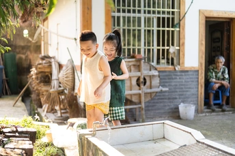 一些小孩子站在一所房子的外面 一个小女孩在一个桶里洗手。