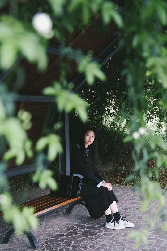 一个穿着黑色连衣裙的年轻女子坐在公园长凳上 树下。
