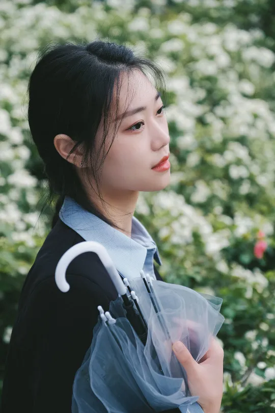 一位穿着黑色连衣裙的年轻女子手持一把透明的伞在白色花朵前。