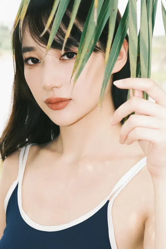 一位绿眼睛的年轻女子用手遮住脸庞 背后有一棵棕榈树。