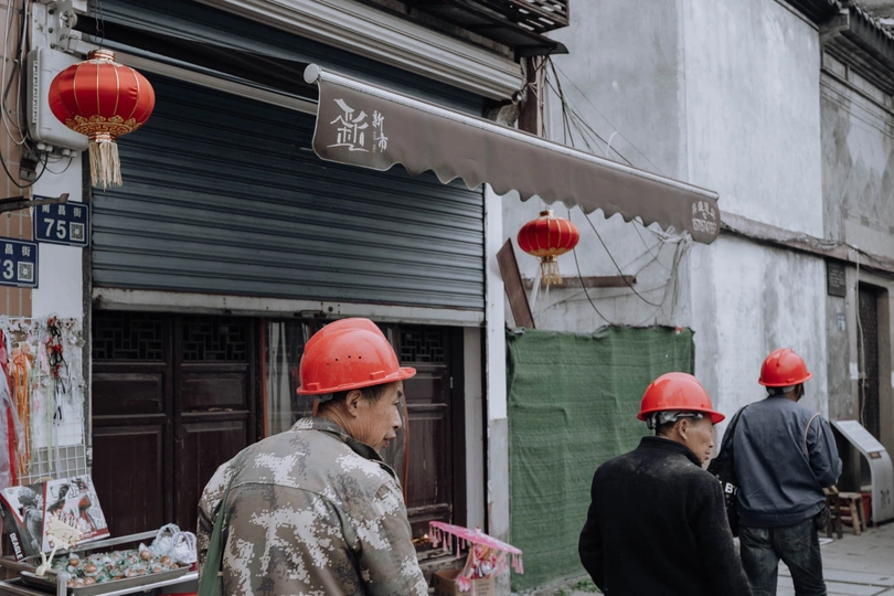 一名戴着红色安全帽的男子走在靠近建筑物的人行道上 旁边有推车的一名工人。