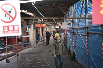 一个戴安全帽的男人走在靠近脚手架和标志的人行道上。