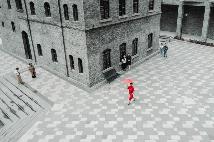 一个人穿着红色连衣裙 拿着红色雨伞 在两个大型建筑之间的庭院中行走。