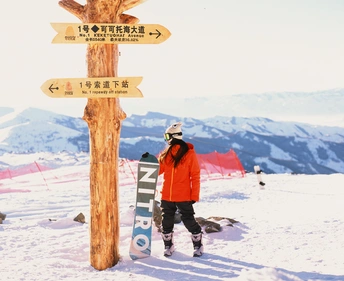 一位雪板运动员站在一个白雪皑皑的山顶旁边 旁边有一根木杆 上面挂着一块牌子。