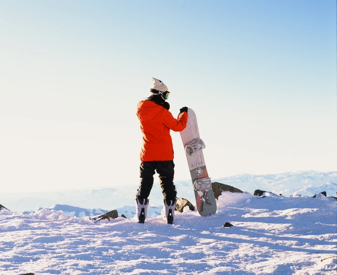 一个穿着橙色夹克的男人站在雪山上 手里拿着滑雪板。