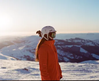 一位穿着橙色夹克和头盔站在雪地上的女人 正在注视着滑雪者。