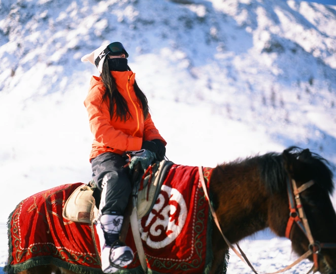 一位穿着橙色夹克的女骑手骑着一匹棕色的马 背景是雪山。