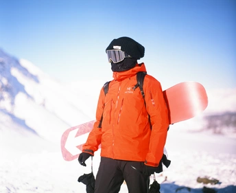 一个穿着橙色夹克的雪板运动员 手拿红色雪板 沿着一个雪地山坡向下走 背景是座山。