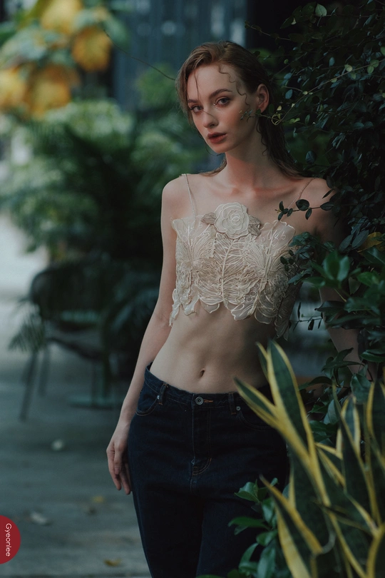 一位穿着衬衫和牛仔裤的年轻女子站在人行道旁靠近灌木丛和植物。