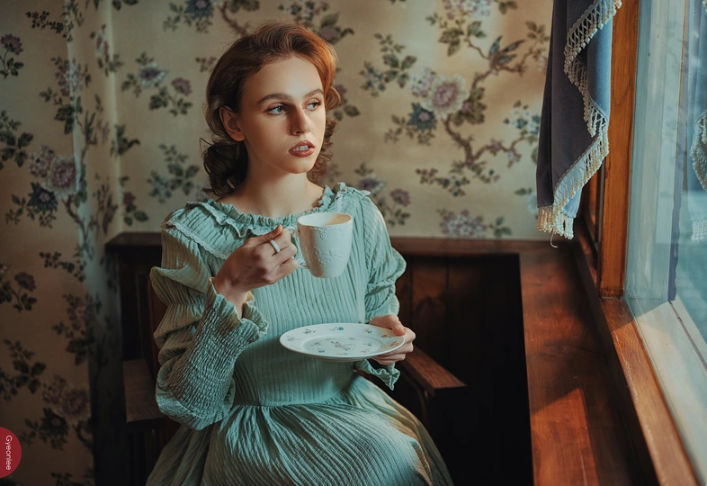 一个穿着绿色连衣裙的年轻女子坐在桌子旁 手里拿着一杯茶或咖啡 正看着窗外一个穿着蓝色连衣裙的年轻女子