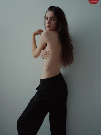 一位穿着黑色裤子和无上衣的年轻女子站在灰色墙壁前
