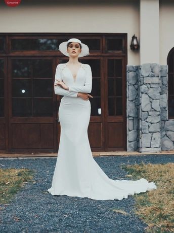 一位穿着白色连衣裙带拖尾的女人在石头房子前戴着帽子穿着长袍站立着