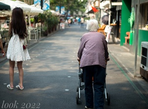 一个老人推着一个助行器走在人行道上 旁边是一个走在街上的小女孩