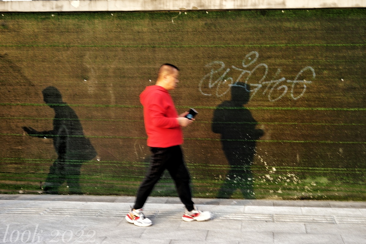 一个穿着红色衬衫的男人走在人行道上 墙上有涂鸦和他的影子
