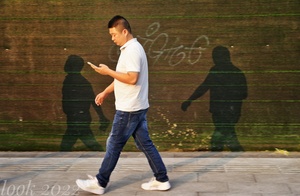 一个年轻人走在人行道上看手机 身后的墙上有他的影子