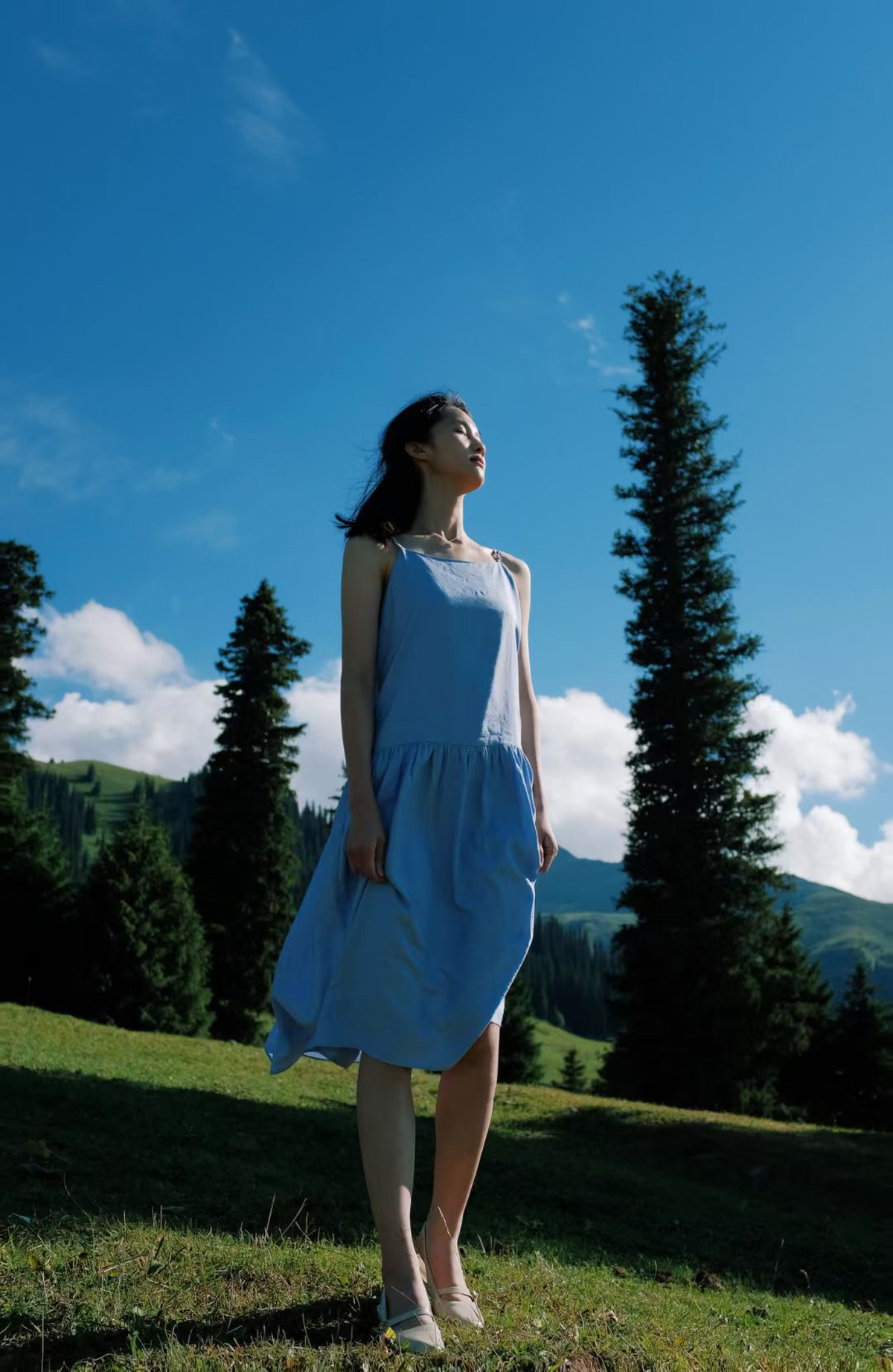 一位穿着蓝色连衣裙的年轻女子在有树木和山脉背景的田野中行走