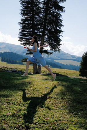 一位穿着蓝色连衣裙的年轻女子跑过一片草地上长满树木和山脉背景的草场 而她的影子在飞盘上。