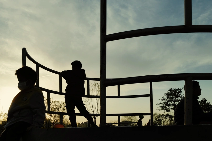 一个年轻男孩站在日落时分的窗户前 背景中有人玩耍。