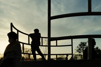 一个年轻男孩站在日落时分的窗户前 背景中有人玩耍。