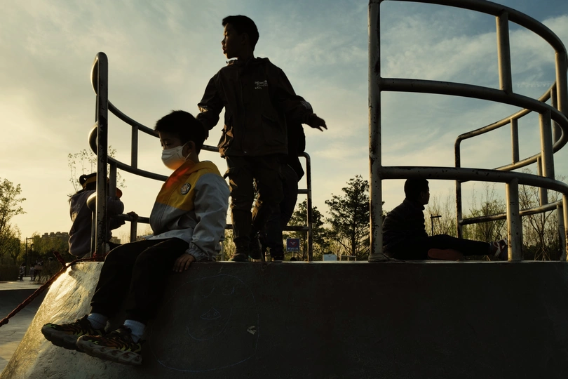 一个戴面罩的年轻男孩在滑板公园骑滑板 周围有其他人站立。