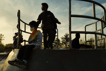 一个戴面罩的年轻男孩在滑板公园骑滑板 周围有其他人站立。