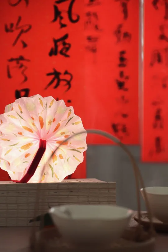 一张桌子 上面放着一盏灯、一个碗和一个花瓶 放在一堵红色墙壁前 墙壁上写着字。