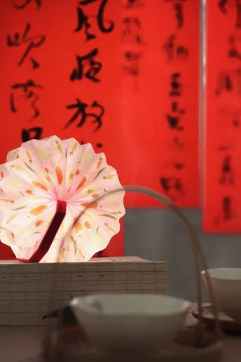 一张桌子 上面放着一盏灯、一个碗和一个花瓶 放在一堵红色墙壁前 墙壁上写着字。