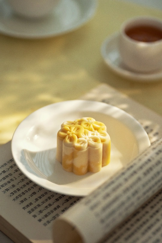 一个小甜点放在一张白色的盘子上 盘子旁边放着一杯茶和一本书。