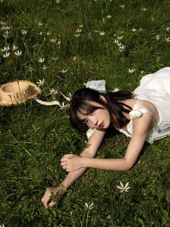一个穿着白色裙子的年轻女子躺在绿草地上 周围是野花 一只猫在附近躺着。