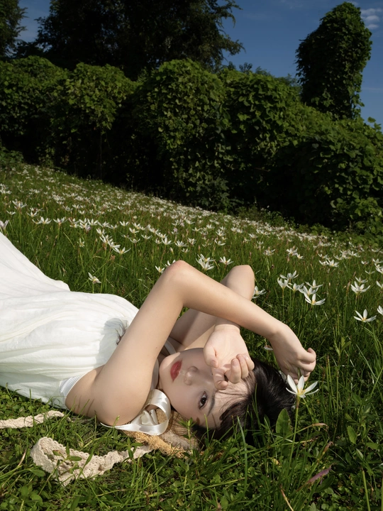 一位穿着白色连衣裙的年轻女子躺在草地上的花丛中。