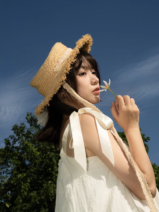 一位穿着白色连衣裙和草帽的年轻女子 背景是蓝天和一朵花。