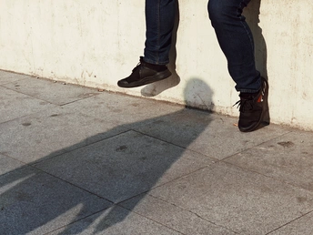 一个穿着牛仔裤和滑板的男人靠在墙上 他的影子落在人行道上。