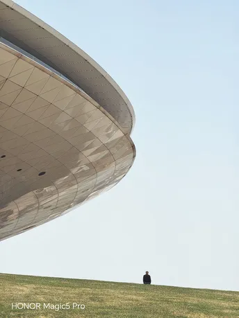 一个人坐在绿草丛生的小山丘上 背景有一座大型建筑。